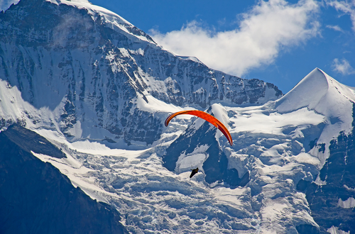 Paragleiter mit roten Schirm. Dahinter schneebedckte Felswände eines Gebirges